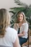 Kelley Nay chats with a client in a warmly lit room. Kelley sits on a tan leather chair, wearing glasses and a cream sweater, appearing attentive and approachable. Green plants and soft furnishings create a welcoming, comfortable atmosphere.